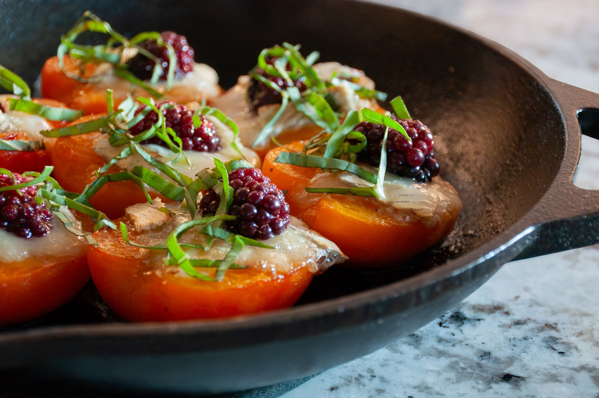 cast iron skillet with broiled apricots and brie garnished with blackberries and slivered basil